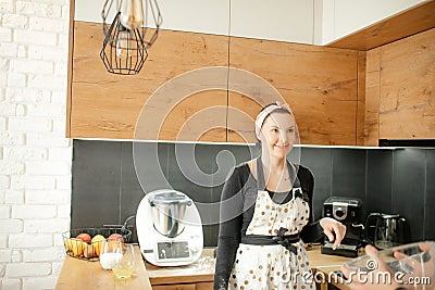 Portrait of young smiling beauteous woman wearing dirty white apron with dots, grey sweatshirt, standing in kitchen. Stock Photo