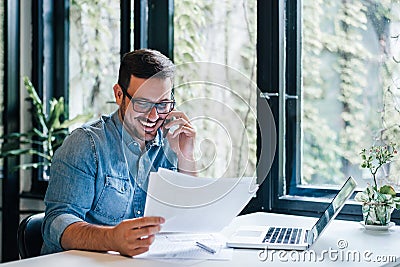 Portrait of young smiling cheerful entrepreneur in casual office making phone call while working with charts and graphs looking at Stock Photo