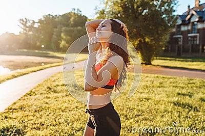 Portrait of young sporty woman in black and pink top and black shorts standing on green lawn at sunrise in sunny day. Stock Photo