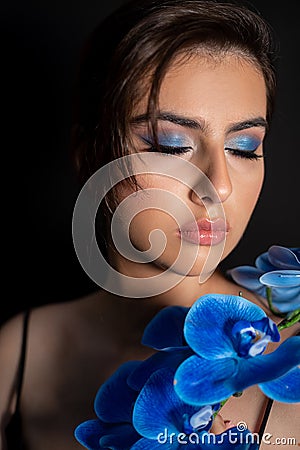 Portrait of young stunning woman with closed eyes, bright blue eyeshadows covering shoulder with branch of blue orchid. Stock Photo