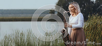 Portrait of young woman playing saxophone on bank of the river in reeds, girl with woodwind musical instrument on nature Stock Photo