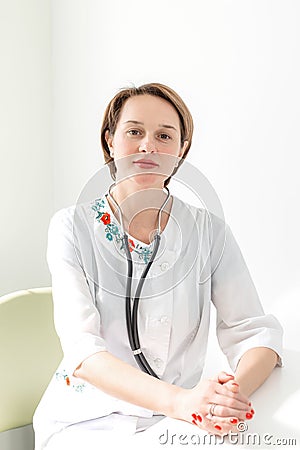 Portrait of a young woman professional doctor with short haircut, in a bright medical room, in a white coat with a phonoscope on Stock Photo