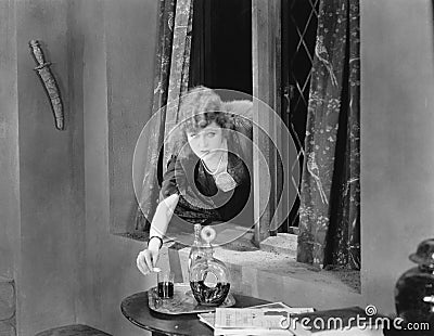 Portrait of a young woman reaching through a window and pouring poison into a glass Stock Photo