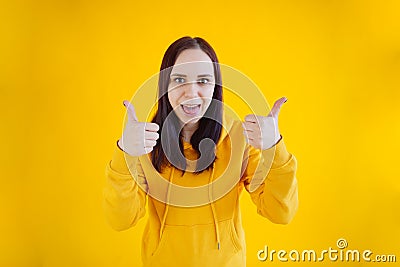 Portrait of young woman smiling and showing thumbs up on yellow background. Close up of happy brunette in yellow hoodie Stock Photo