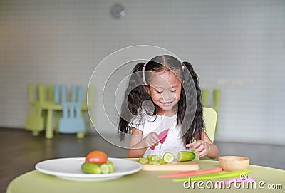 Portraits of happy Asian child girl slicing cucumber vegetable on chopping board at play room. Kid play chef cooking Stock Photo