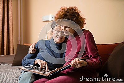 Positive aged ladies looking album photos sitting sofa at home, cheerful friends. Senior woman and her mature nurse watching photo Stock Photo
