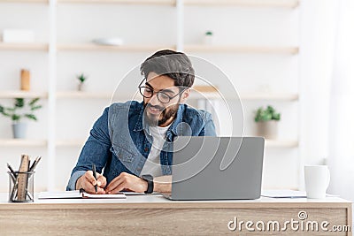 Positive arab male manager working from home during COVID-19 pandemic, using modern laptop and taking notes Stock Photo