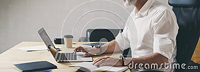 Positive bearded businessman using mobile laptop computer while sitting at wooden table at modern coworking place. Wide Stock Photo