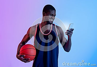 Positive black basketball fan watching championship online, cheering for his favorite team on smartphone in neon light Stock Photo