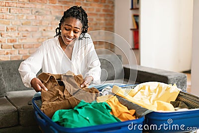 Positive black woman packing suitcase for vacation, sitting near open suitcase and preparing for travel at home Stock Photo