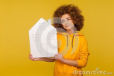 Positive bright woman with fluffy curly hairstyle in hoody looking at paper house with thoughtful expression Stock Photo