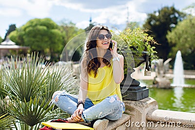 Positive brunette girl is speaking on phone in summer park. She is sitting on fence, smiling to side Stock Photo