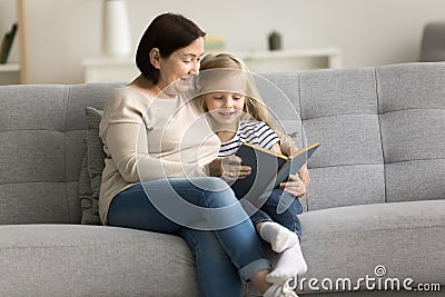 Positive grandma reading book to granddaughter kid on comfortable couch Stock Photo