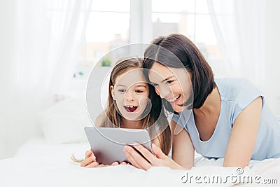 Positive cheerful small female kid and her young mother watch interesting cartoon on tablet, connected to wireless internet, pose Stock Photo