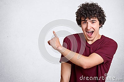 Positive curly guy keeps mouth opened, exclaims with happiness, points aside at blank copy space for your advertising content, wea Stock Photo