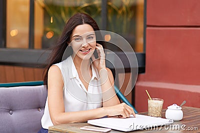 Positive dark haired woman in white outfit, surrounded with modern gadgets, sits at coffee shop, smiles gently at camera, being in Stock Photo