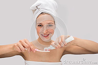 Positive freckled young woman looks on camera and smiles. She puts some toothpaste on tooth brush. Model has white Stock Photo