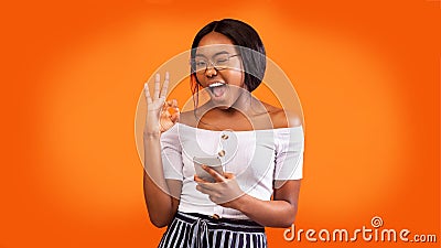 Positive Girl Holding Phone Gesturing Okay And Winking, Orange Background Stock Photo