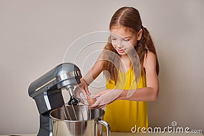 A positive girl of 9-10 years cooks homemade cake in the kitchen, beats eggs in a mixer on the kitchen table. Against Stock Photo