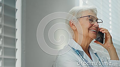 Positive grey haired mature lady doctor with glasses talks on mobile phone with family member near window Stock Photo