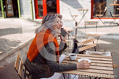 Positive grey haired senior lady talks on phone at table on outdoors cafe terrace Stock Photo