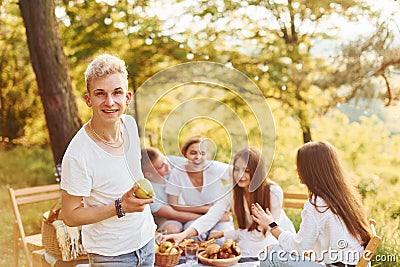 Positive guy with apple standing in front of people. Group of young people have vacation outdoors in the forest. Conception of Stock Photo