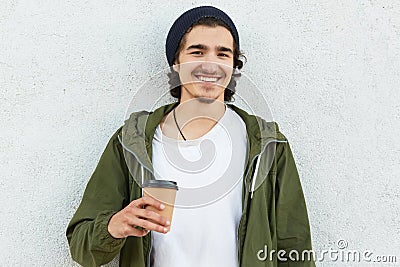 Positive hipster in stylish black headgear, white t shirt and anorak, holds paper cup of coffee, enjoys spare time, isolated over Stock Photo