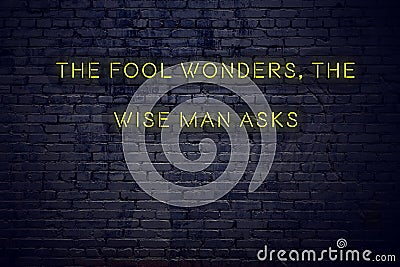 Positive inspiring quote on neon sign against brick wall the fool wonders the wise man asks Stock Photo