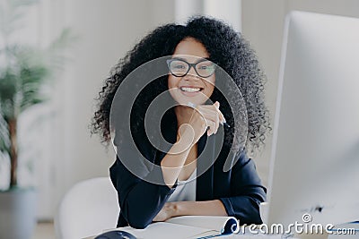 Positive international female student with crisp hair, wears transparent glasses, holds pen in hand, makes accountings, sits in Stock Photo