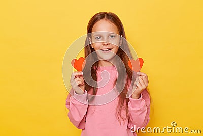Positive joyful brown haired little girl wearing pink sweatshirt isolated over yellow background showing two little red hearts Stock Photo