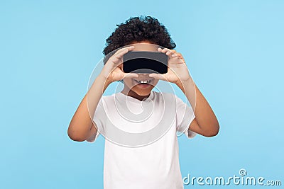 Positive lovely happy little boy with curly hair covering eyes with cellphone and smiling Stock Photo