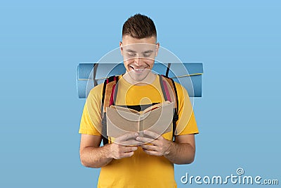 Positive millennial hipster with camping gear reading dictionary or guidebook over blue studio background Stock Photo