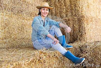 Positive relaxed asian female farmer resting on hayloft after work at farm, sitting on straw stack and smiling Stock Photo