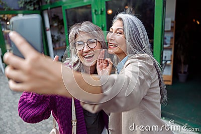 Positive senior Asian lady with grey haired friend take selfie on modern city street Stock Photo