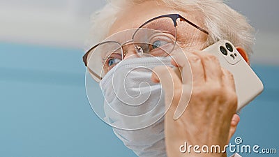 Positive senior lady doctor in disposable mask talks on mobile phone consulting patient in hospital office Stock Photo