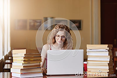 Positive tender sweet girl with curly hair working on her laptop being in reading hall with huge bunch of books on desk, focused Stock Photo