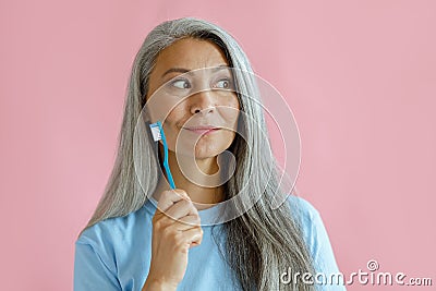 Positive thoughtful middle aged Asian woman holds toothbrush in studio Stock Photo