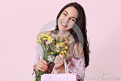 Positive young brunette European woman with happy expression, tilts head, holds gift bag and pretty flowers, has sincere tender Stock Photo