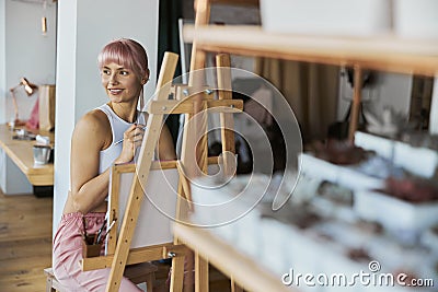 Positive young female model with pink hair sits at wooden easel with canvas Stock Photo