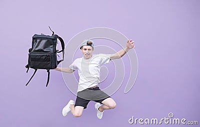 Positive young man in a white T-shirt jumping with a backpack against the background of a purple wall Stock Photo