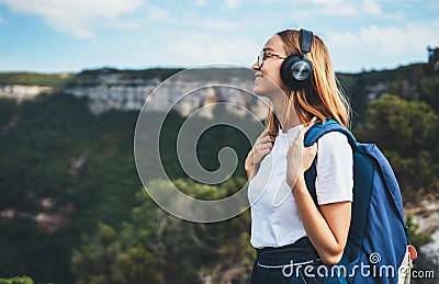 Positive young traveler girl with headphones and backpack stands on top of mountain, active tourist woman hiking enjoys beautiful Stock Photo