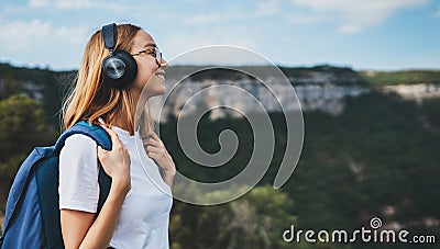 Positive young traveler girl with headphones and backpack stands on top of mountain and enjoys beautiful landscape, active tourist Stock Photo