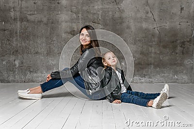 Positive young woman and little girl in similar rock style clothing jeans and black leather jackets sitting back to back Stock Photo