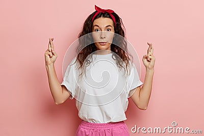 Positive young woman, making a wish, she makes finger crossed and happily looking into the camera, isolated over pink Stock Photo