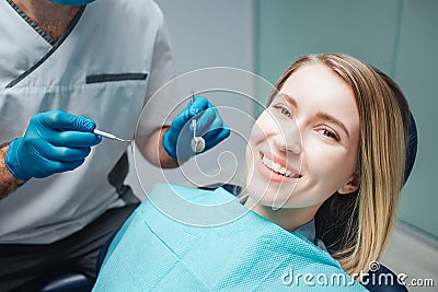 Positive young woman sit in chairin dentistry and smile to camera. She show beautiful smile. Doctor hold tools for teeth Stock Photo