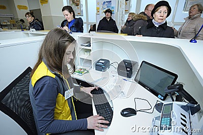 At the post office, reception: postal worker sitting at the desk set with computer and terminals, visitors waiting in front of a Editorial Stock Photo