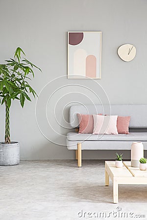 Poster and clock above settee with pink pillows in grey flat interior with plant and table. Real photo Stock Photo