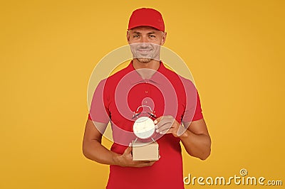 Postman delivery worker. Handsome man red cap hold alarm clock yellow background. Delivering purchase. Open hours. Time Stock Photo