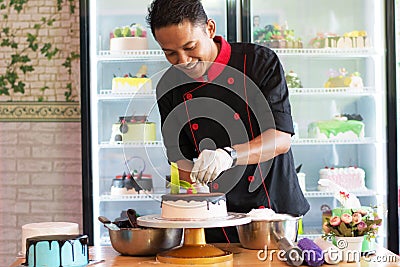 Potrait of asian male pastry chef in a black uniform decorating round vanila cake with melted chocolate and red cherry Stock Photo