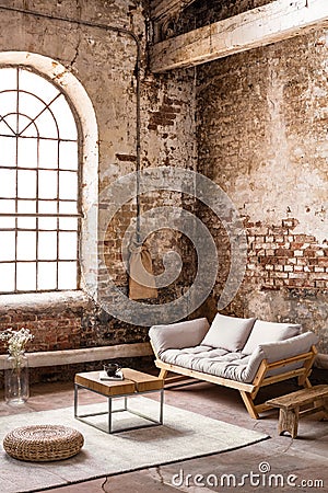 Pouf and table on rug in front of wooden sofa in spacious loft interior with window Stock Photo